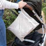 A person pushes a stroller with a Bumkins Wet/Dry Bag: Wander attached to the handle amid lush green foliage in the background.