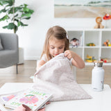 A young girl reaches into a Bumkins Wet/Dry Bag: Wander at a white table with books and water bottle in a bright, plant-filled room.