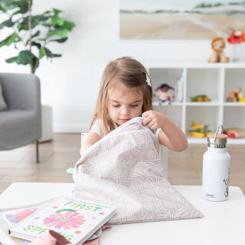 A young girl reaches into a Bumkins Wet/Dry Bag: Wander at a white table with books and water bottle in a bright, plant-filled room.