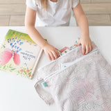 A child fills a Bumkins Wet/Dry Bag: Wander with treasures, while a colorful book sits nearby on a white table.
