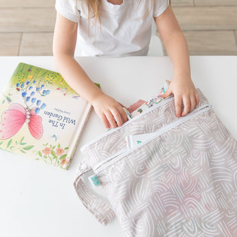 A child fills a Bumkins Wet/Dry Bag: Wander with treasures, while a colorful book sits nearby on a white table.