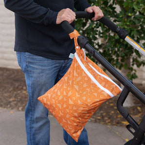 A person strolls outdoors with a Bumkins Wet/Dry Bag: Grounded, featuring zippered compartments, hanging from the stroller handle.