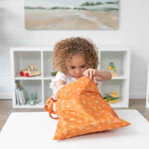 A curly-haired child peers into a Bumkins Wet/Dry Bag: Grounded at a white table, surrounded by toy-filled shelves and artwork.