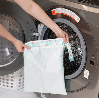 A person holds a bag from Bumkins Wet Bag + Wet/Dry Bag Set: Rainbow in front of an open front-loading washing machine.