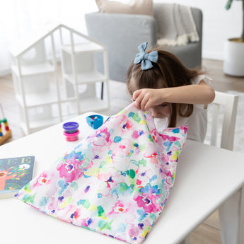 A young girl with a blue bow looks into her Bumkins Wet Bag + Wet/Dry Bag Set: Watercolor at a white table in a cozy room.