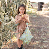A girl smiles in front of cornstalks, holding a bag from Bumkins Wet Bag + Wet/Dry Bag Set: Camp Gear on her arm and a corn cob in her hands.