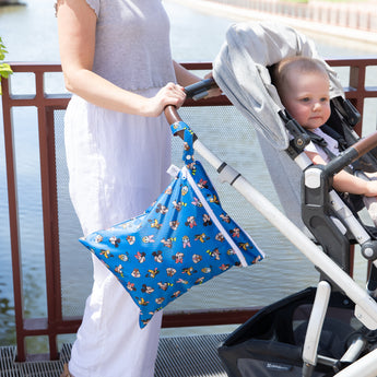 An adult pushes a stroller with a child near a waterfront with Bumkins Wet/Dry Bag: Mickey & Friends Forever attached to the handle.