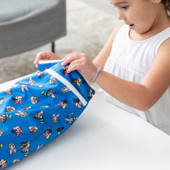 A child in a white dress opens a blue Bumkins Wet/Dry Bag: Mickey & Friends Forever while sitting at a white table.
