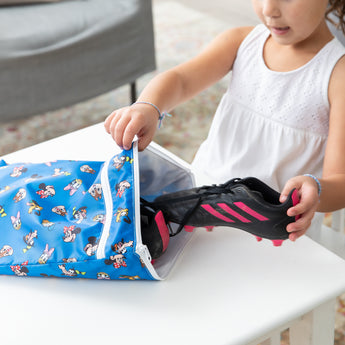 A child places a soccer cleat into a blue Bumkins Wet/Dry Bag: Mickey & Friends Forever on a white table.