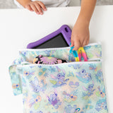 A child puts colorful pens and a purple tablet into the Bumkins Wet/Dry Bag Set: Aloha Stitch on a white table.