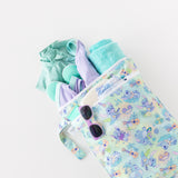 A bag from Bumkins Wet Bag + Wet/Dry Bag Set: Aloha Stitch lays on a white background with a towel, sunglasses, and swimwear peeking out.