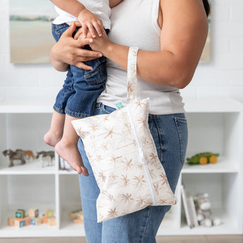 A woman holds a baby and a bag from Bumkins The Chaos Coordinator Deluxe 5-Piece Set: Palm Daze on her arm in front of toy-filled shelves.