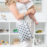 A woman holds a baby and a bag from Bumkins On-The-Go Mealtime 6-Piece Set: Charcoal Check + Grey in front of toy-filled shelves.