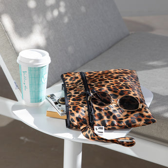 A Roamio by Bumkins Wet/Dry Clutch: Leopard with sunglasses & a camera sits on a white table with a coffee cup by a gray chair outdoors.