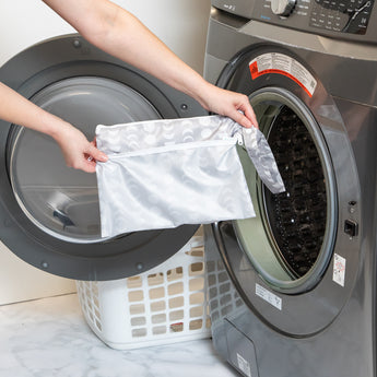 A person holds a bag from Bumkins The Chaos Coordinator Deluxe 5-Piece Set: Lunar Phase in front of an open front-loading washing machine.