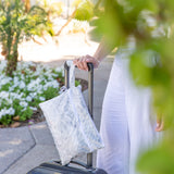 A person in white pants places their hand on a suitcase handle with a Roamio by Bumkins Wet/Dry Clutch: Disco Check, surrounded by greenery.