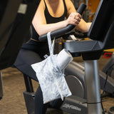 A person uses a stationery bike at a gym, with a Roamio by Bumkins Wet/Dry Clutch: Disco Check holding a towel hanging on the handle.