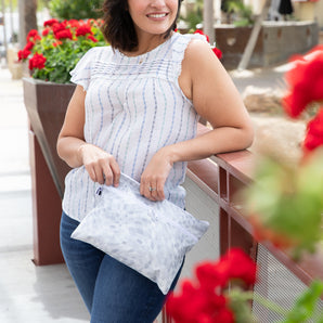 A woman in a striped blouse holds a Roamio by Bumkins Wet/Dry Clutch: Disco Check, smiling by red flowers while leaning on a railing.