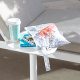 A Roamio by Bumkins Wet/Dry Clutch: Disco Check with pink sunglasses and books sits by a coffee cup on a table next to a gray cushioned chair.