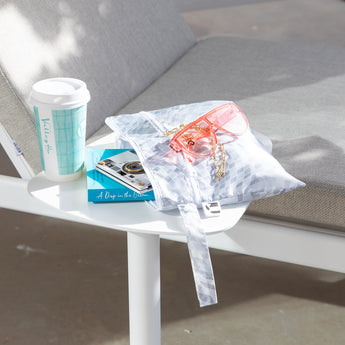 A Roamio by Bumkins Wet/Dry Clutch: Disco Check with pink sunglasses and books sits by a coffee cup on a table next to a gray cushioned chair.