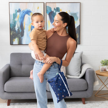 A smiling woman holds a toddler in a living room with paintings and a gray couch; she's carrying Bumkins Wet/Dry Clutch: Evil Eye on her arm.