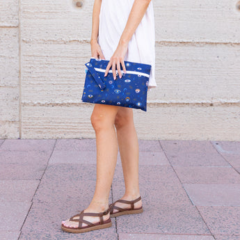 Someone in a light dress holds a Bumkins Wet/Dry Clutch: Evil Eye on tiled pavement against a plain wall.