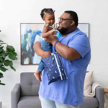 A smiling man in a blue shirt holds a young child and a Bumkins Wet/Dry Clutch: Evil Eye in a modern living room with artwork.