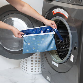 A person holds Bumkins Wet/Dry Clutch: Evil Eye in front of a front-loading washing machine.