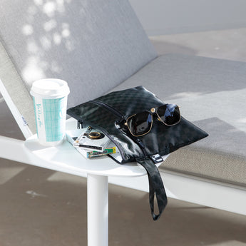 Roamio by Bumkins Wet/Dry Clutch: Rich Black sits on a table outdoors next to a lounge chair with sunglasses, books, and a coffee cup.