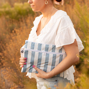 A woman in white blouse holds a Roamio by Bumkins Wet/Dry Clutch: Nautical Stripe among dry grass outdoors in golden sunlight.