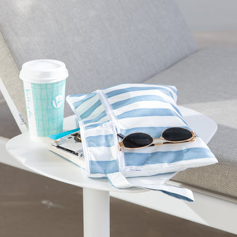 A Roamio by Bumkins Wet/Dry Clutch: Nautical Stripe with sunglasses & books sits on a table next to a coffee cup by a gray cushioned chair.