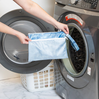 A person holds a bag from Roamio by Bumkins Wet Bag + Wet/Dry Clutch Set: Denim On Denim in front of an open front-loading washing machine.