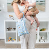 A mom holds a baby and a bag from Bumkins On-The-Go Mealtime 5-Piece Set: Mickey + Minnie Mouse Sunny Side Up Blue by a shelf with toys.