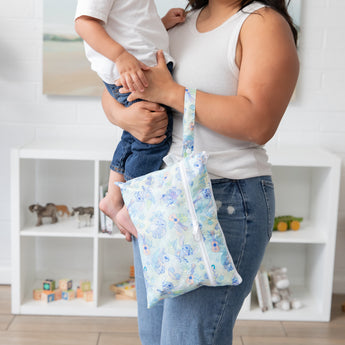 A woman holds a baby and a bag from Bumkins The Chaos Coordinator Deluxe 5-Piece Set: Disney Aloha Stitch front of toy-filled shelves.