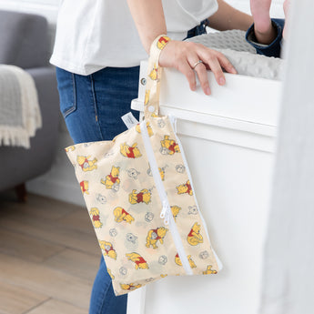 A person in jeans leans against a white changing table with a baby on it; a Bumkins Wet/Dry Clutch: Winnie Loves Hunny hangs from their wrist.