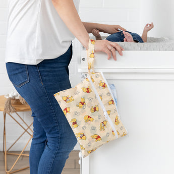 A person stands next to a baby on a changing table, with a Bumkins Wet/Dry Clutch: Winnie Loves Hunny hanging from their wrist.