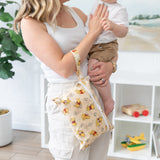 A woman holds a child and a Bumkins Wet/Dry Clutch: Winnie Loves Hunny with plants and toys on a shelf in the background.