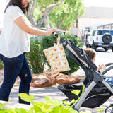 In a sunny setting, a woman in a white shirt and jeans pushes a stroller with a Bumkins Wet/Dry Clutch: Winnie Loves Hunny hanging on the handle.