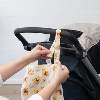 Hands attach a Bumkins Wet/Dry Clutch: Winnie Loves Hunny to a stroller handle against a white brick wall.