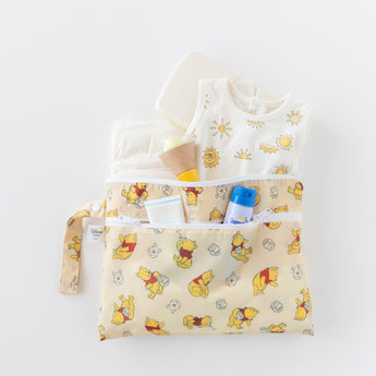 Bumkins Wet/Dry Clutch: Winnie Loves Hunny holds essentials like diapers, wipes, sunscreen, and an outfit on a white background.