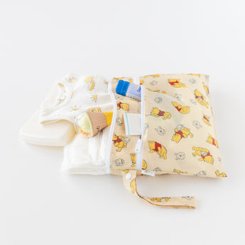 Bumkins Wet/Dry Clutch: Winnie Loves Hunny holds baby essentials on a white background: a printed onesie, wipes, diapers, and cream.