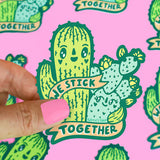 A hand with pink nails holds a Turtle Soup Vinyl Sticker of hugging cacti, banner reads We Stick Together, on a pink background.