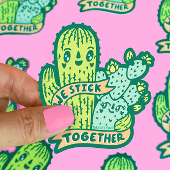A hand with pink nails holds a Turtle Soup Vinyl Sticker of hugging cacti, banner reads We Stick Together, on a pink background.