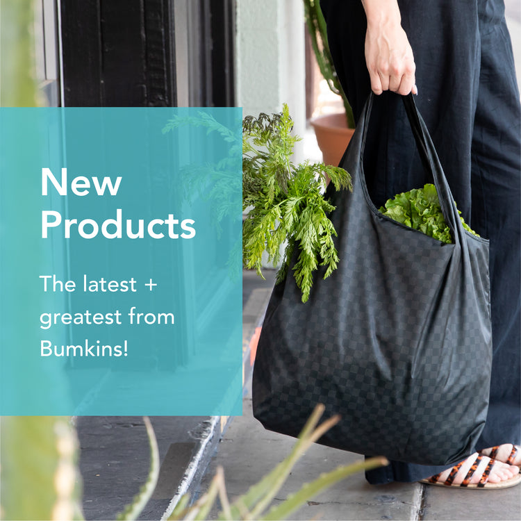 Person holding a checkered reusable bag filled with fresh greens, standing next to a shop entrance. Text on the image reads, New Products: The latest + greatest from Bumkins!.