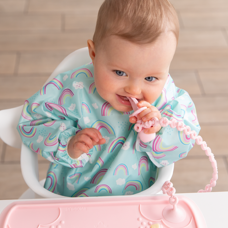 A smiling child in a blue bib with rainbow pattern chews a pink toddler utensil attached to a pink placemat with a pink silicone tether.