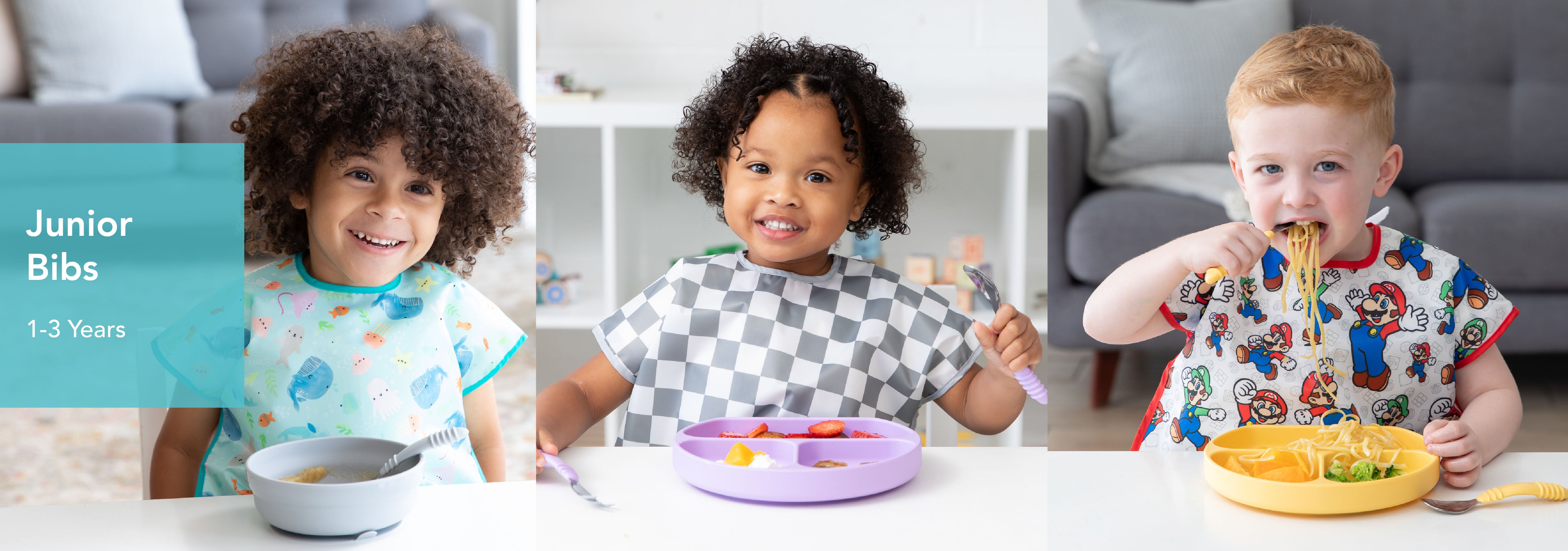 Three toddlers wearing bibs are eating at a table. The first is enjoying cereal with a spoon, the second has a checkered bib and is eating fruit and veggies from a purple plate, and the third is eating spaghetti with a fork, wearing a colorful bib.