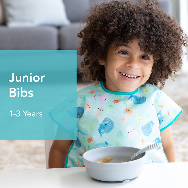 A smiling toddler with curly hair wearing a colorful bib sits at a table with a bowl and spoon. Text on the left reads Junior Bibs 1-3 Years. A couch is in the background.