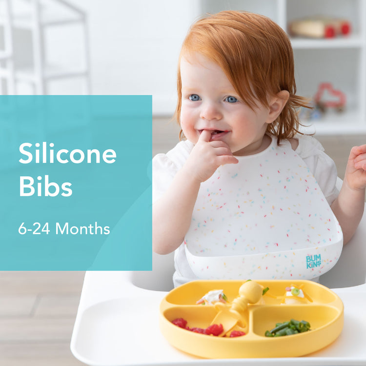 A toddler with red hair sits in a high chair, wearing a speckled silicone bib. They are touching their mouth and smiling. In front of them is a yellow divided plate with food. Text on image reads: Silicone Bibs, 6-24 Months.