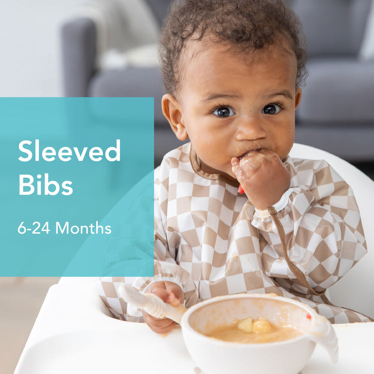 Toddler wearing a checkered long-sleeved bib, eating from a bowl while seated. Text on image reads: Sleeved Bibs 6-24 Months. Background shows part of a gray couch.