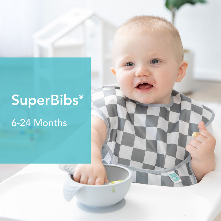 A baby seated in a highchair wears a checkered bib and is touching food in a bowl. Text beside the baby reads, SuperBibs 6-24 Months in a turquoise box. The background is softly lit, showing part of a room.
